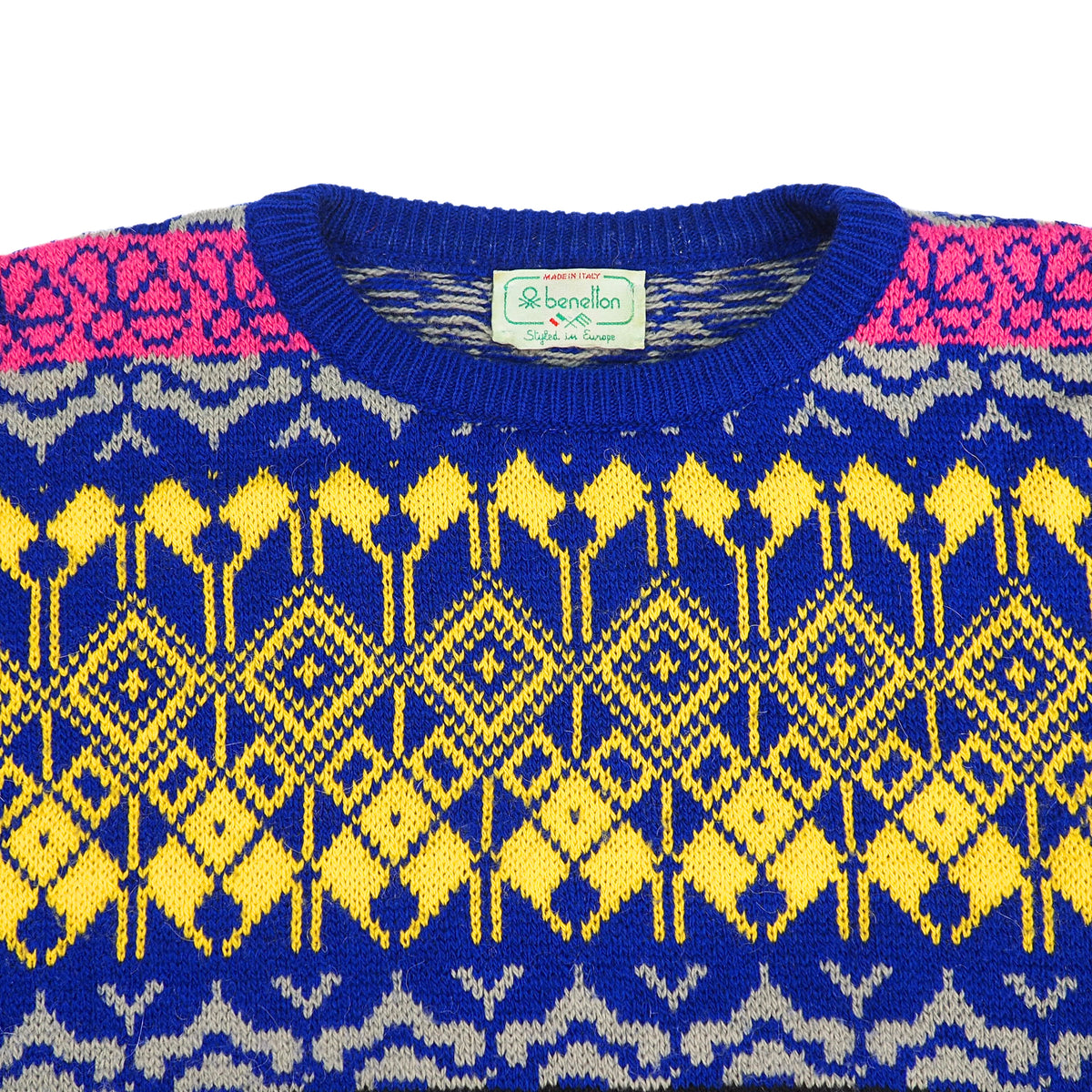 A close-up of a knitted crewneck sweater by Benetton, featuring a vibrant blue base with yellow geometric patterns, pink patterned shoulders, and light gray accents.
