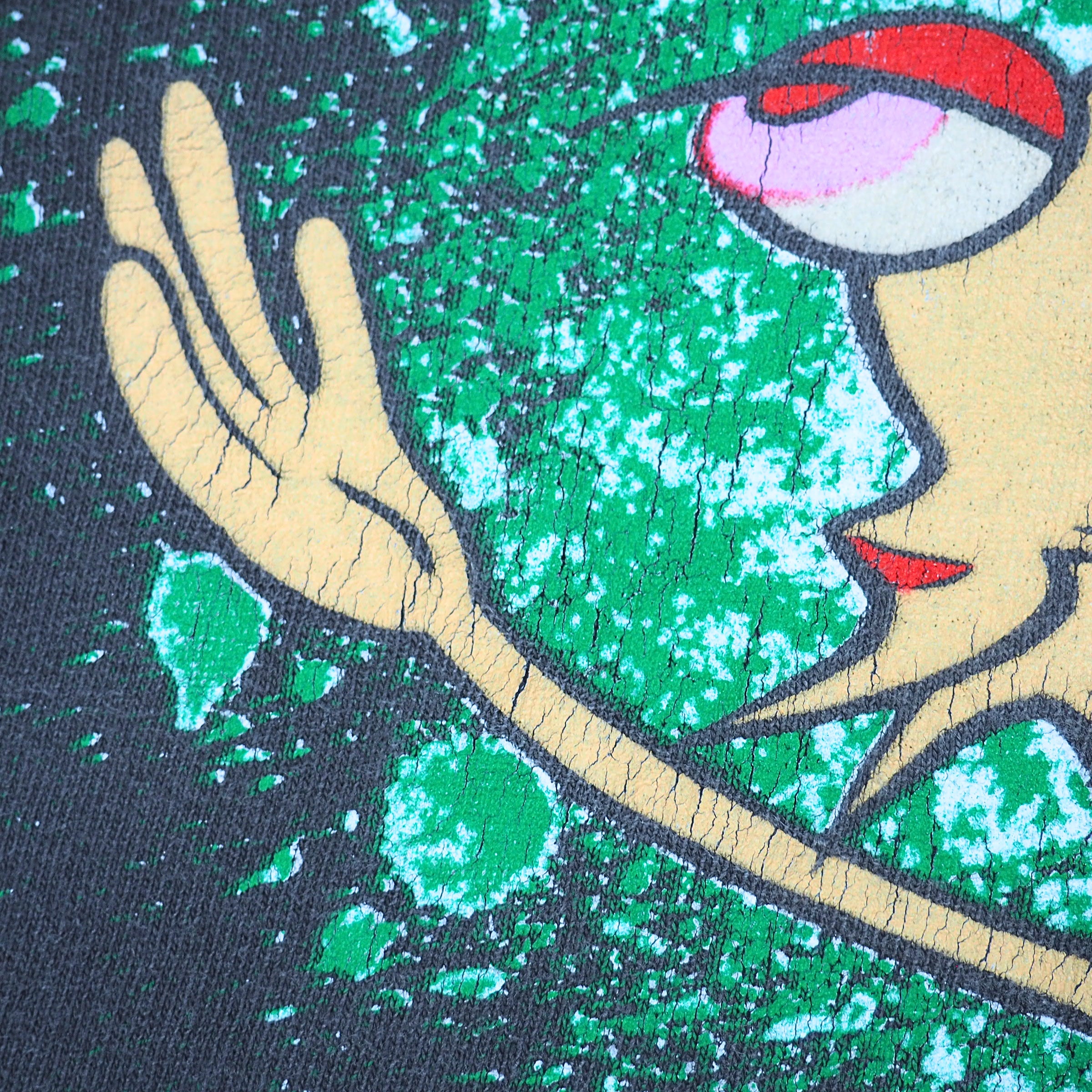 Close-up of a graphic print featuring a stylized face and hand in beige, red, pink, and white, set against a textured green and white splatter background on a dark base.