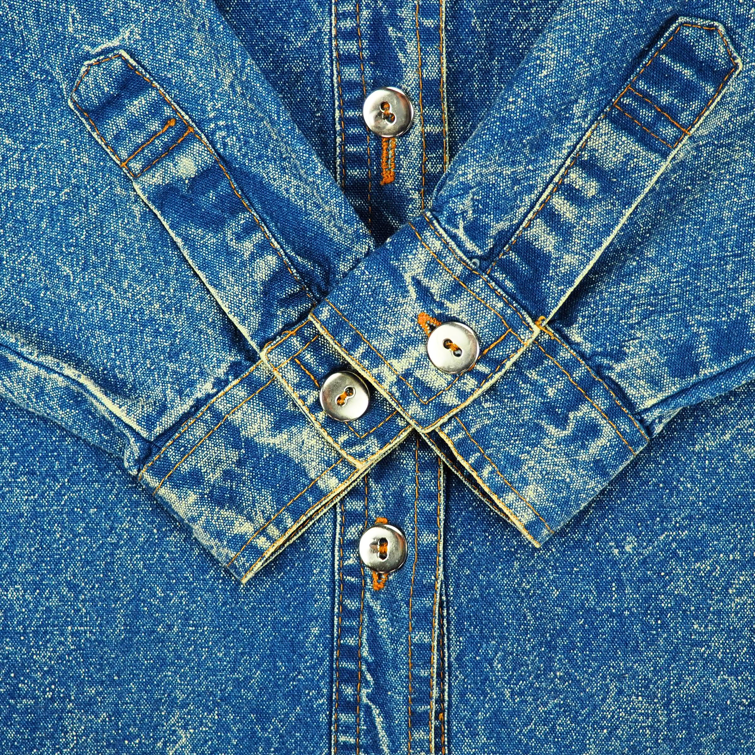 Close-up of a blue acid wash denim sleeve cuffs folded with metallic silver buttons folded onto the body of the shirt