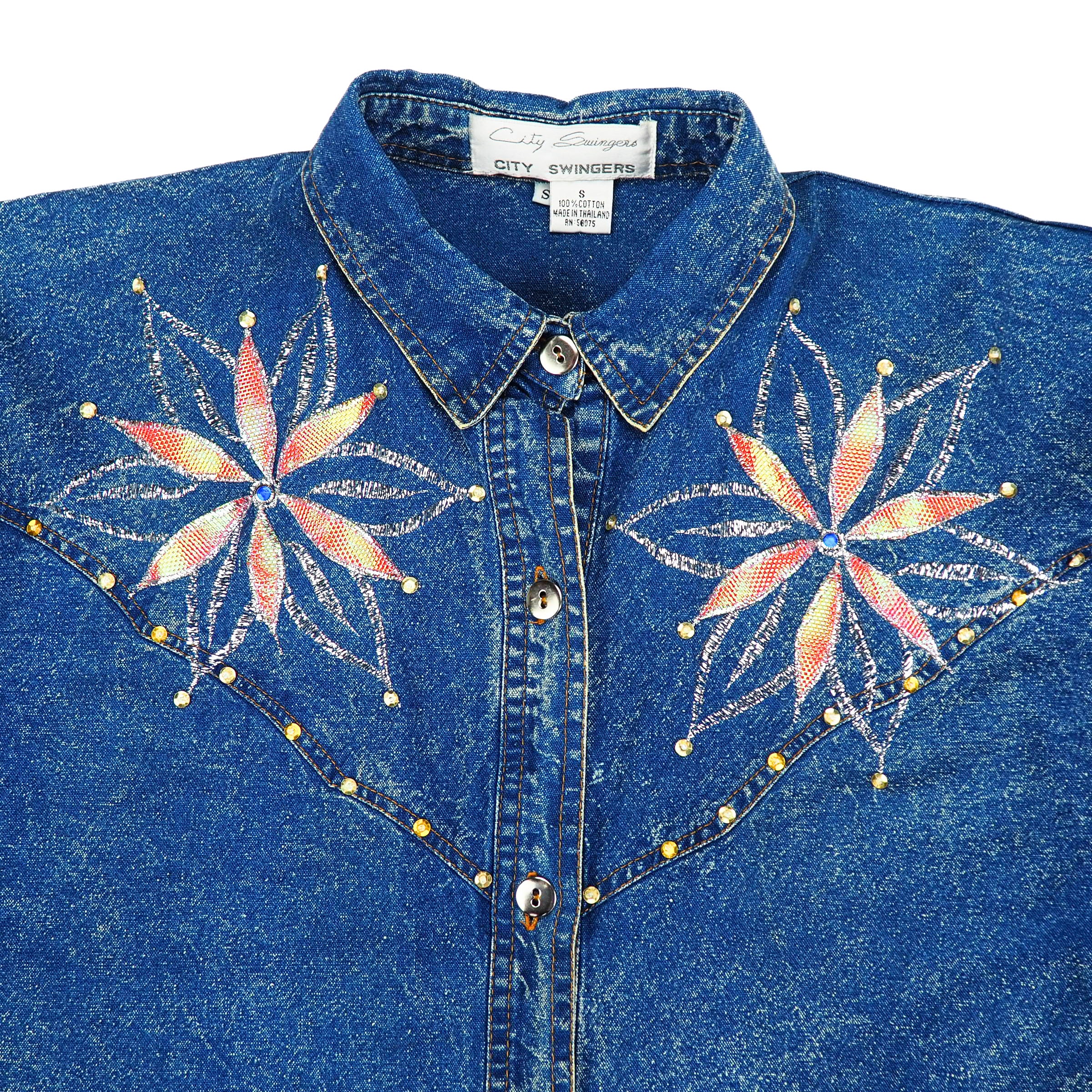 Blue denim shirt with floral embroidery and decorative buttons on a white background