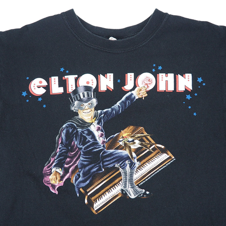 A black short-sleeve graphic t-shirt featuring Elton John's 'My Favorite Thing' design. The graphic shows the text 'ELTON JOHN' in red and white bubble letters, with a cartoon illustration of Elton John in a top hat and tailcoat sitting on a piano and holding a lollipop, surrounded by small blue stars.