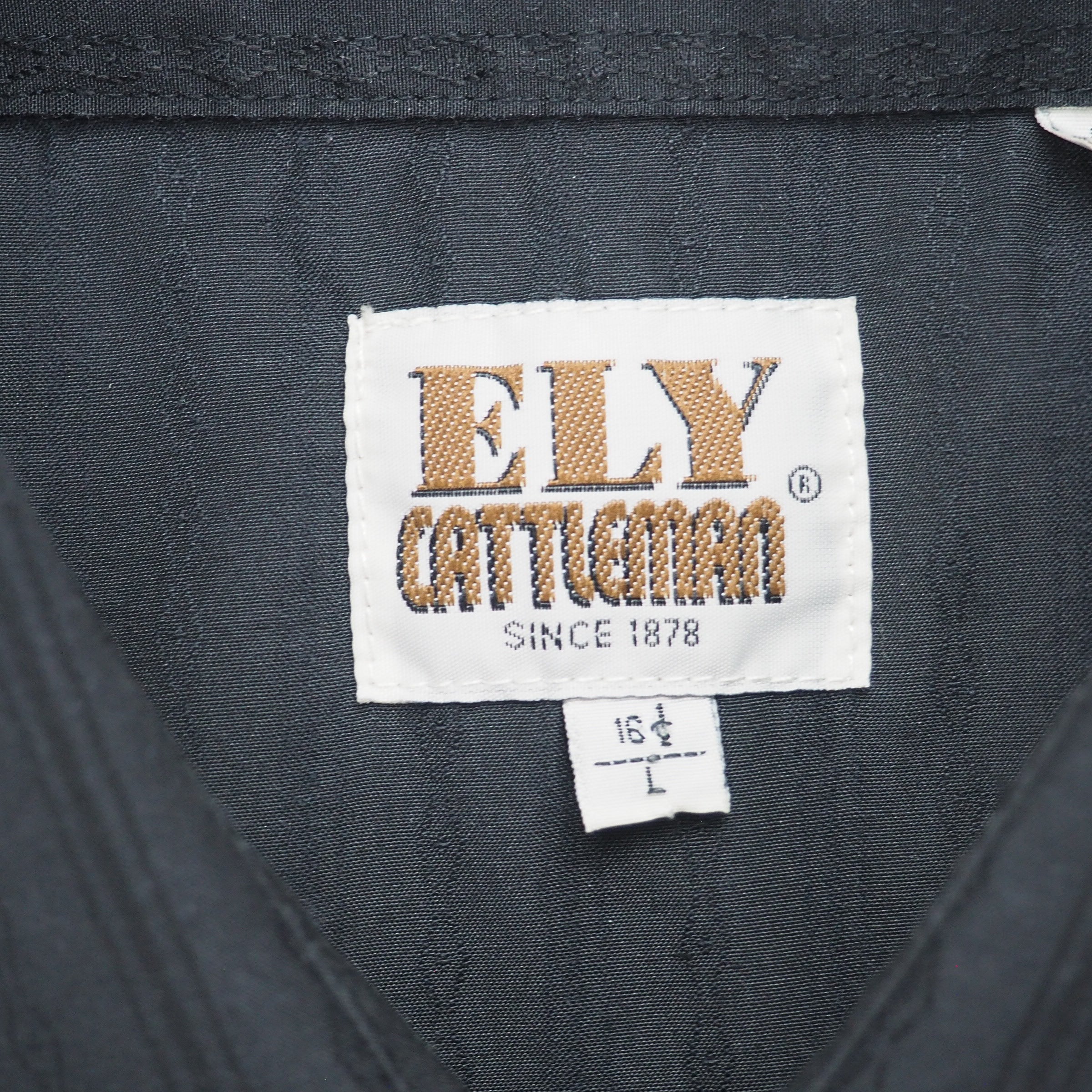 Close up of brand tag reading 'Ely Cattleman Since 1878' on the inside collar of a black shirt.