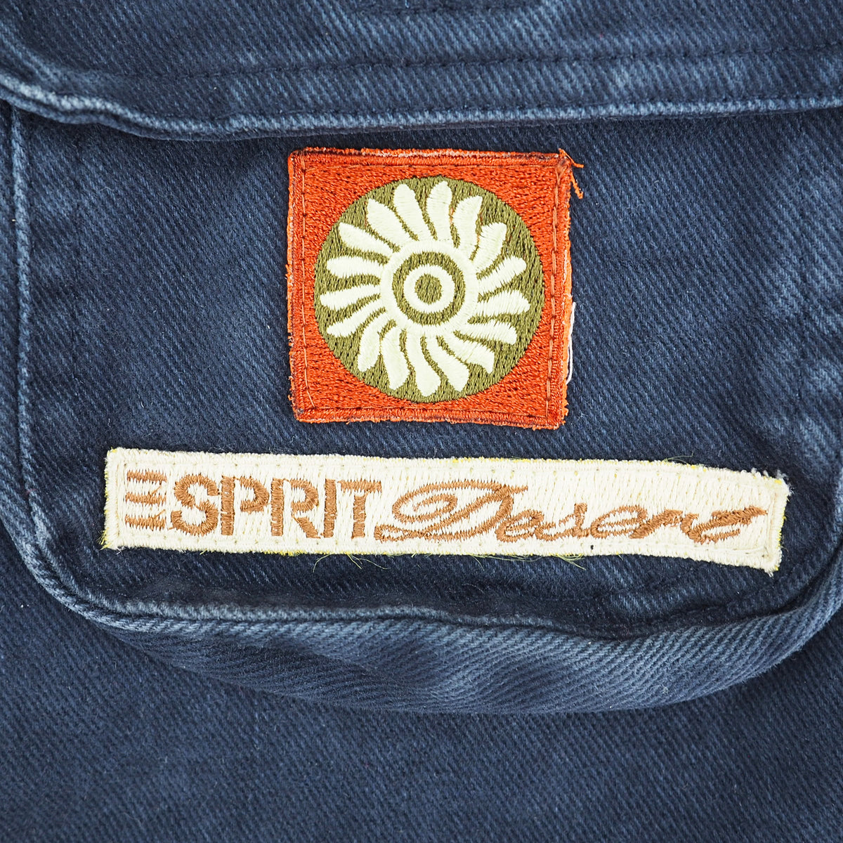 Close-up of a blue denim garment showing two embroidered patches: a square orange patch with a white sunburst motif and a rectangular cream patch reading “ESPRIT DESERT” in orange embroidery.