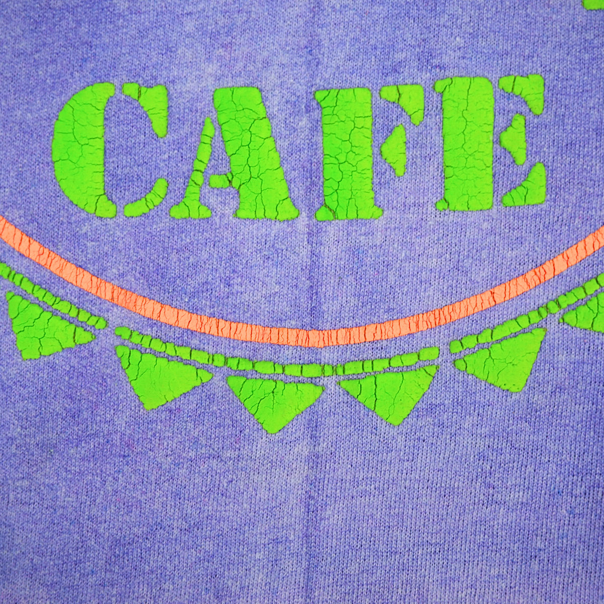 Close-up of a purple knit fabric with bright green puff-print letters spelling "CAFE" and a curved orange line and green arrow heads below.