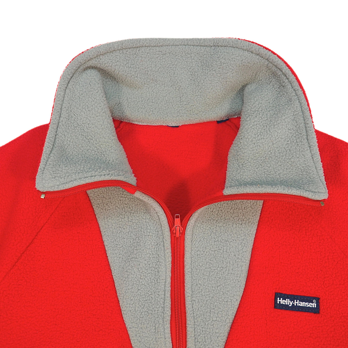 Close-up of a red fleece zip-up jacket with a light gray fleece collar and zipper placket, featuring a small black Helly Hansen logo patch on the left chest.