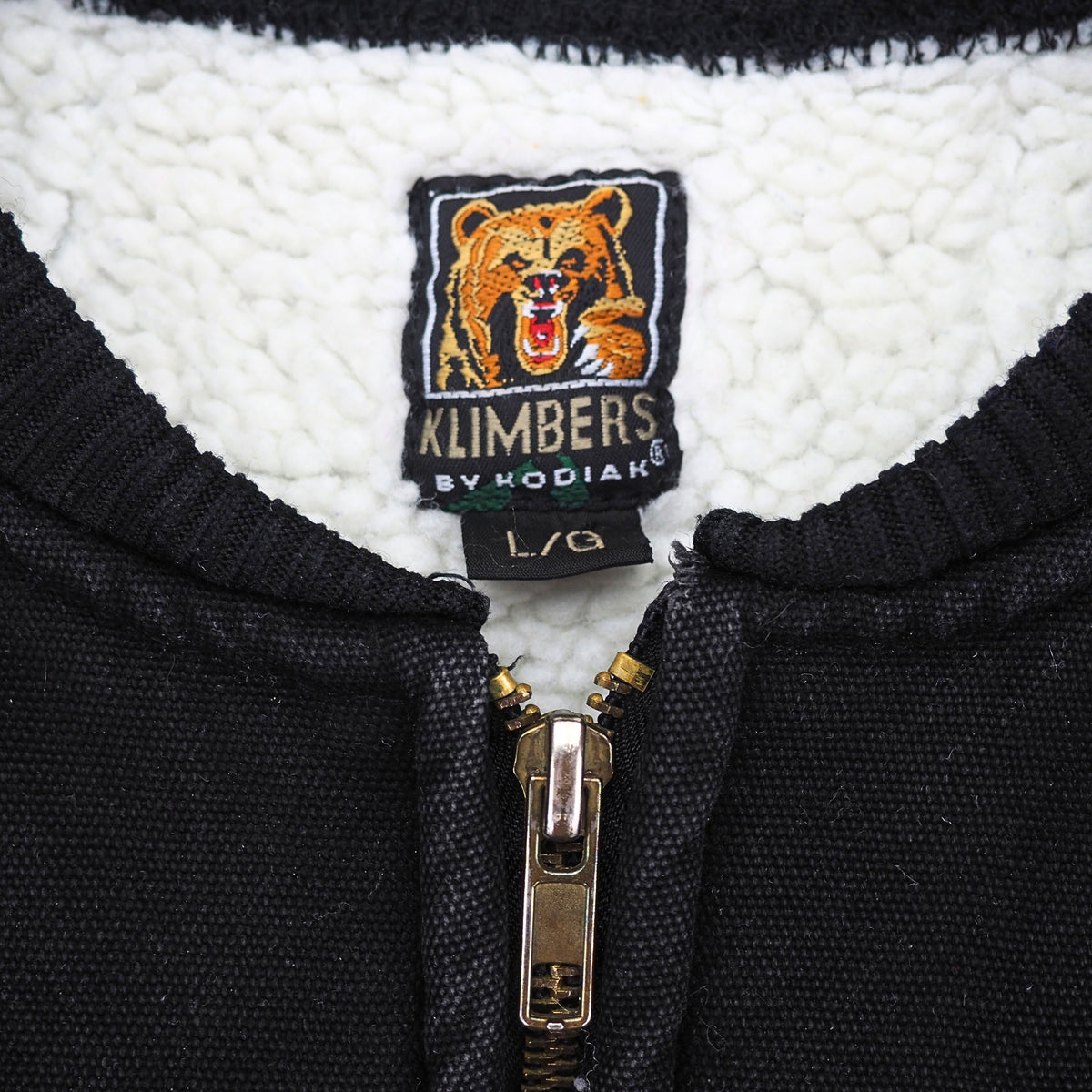 Close up of the brand label 'Klimbers by Kodiak' with a 'L' size tag in the inside collar of a faded black canvas work vest with white sherpa fleece lining.