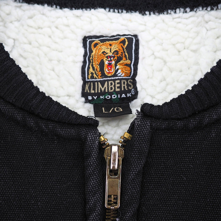 Close up of the brand label 'Klimbers by Kodiak' with a 'L' size tag in the inside collar of a faded black canvas work vest with white sherpa fleece lining.
