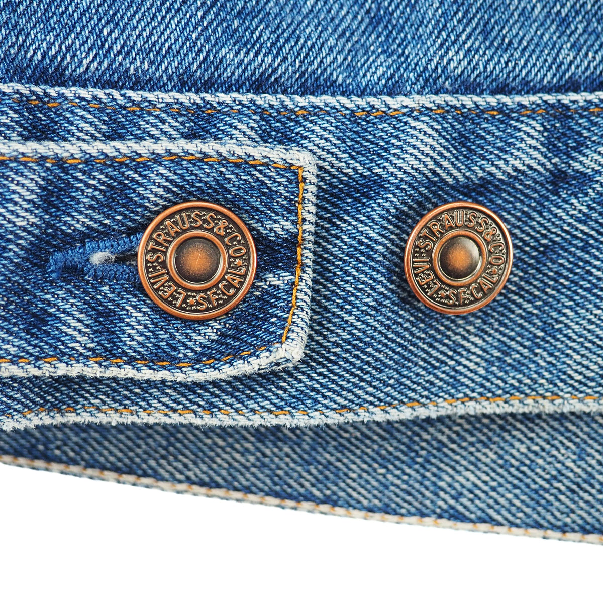 Close-up detail of a blue denim garment showing two round copper-toned snap buttons on the pocket flap, with visible denim texture and contrast stitching.