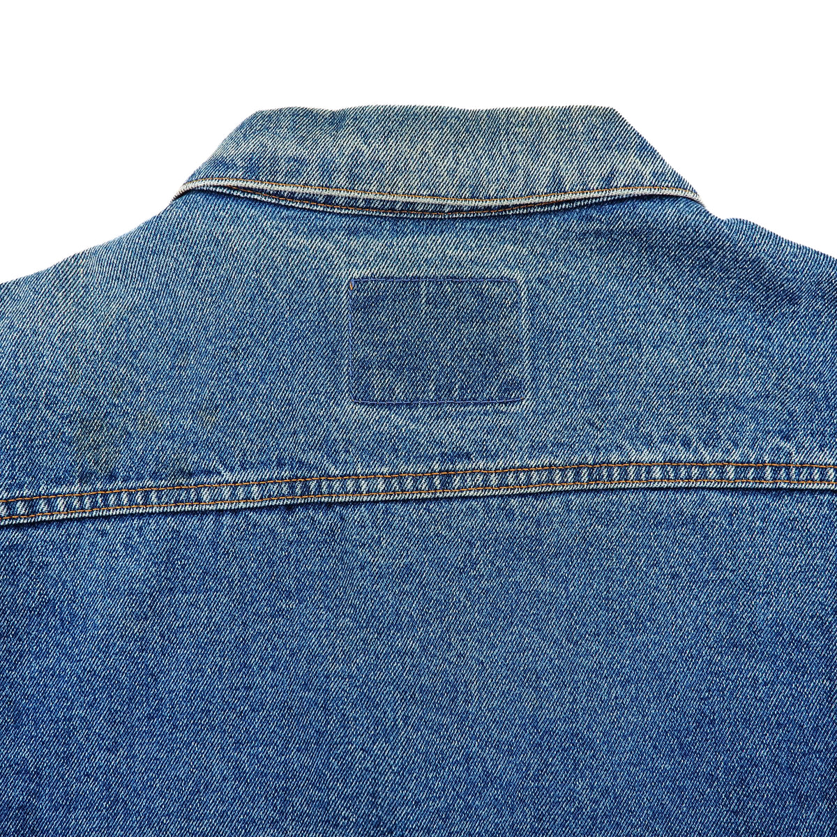 Close-up back view of a blue denim jacket showing the collar, back yoke, seam stitching, and a blank rectangular patch on a white background.
