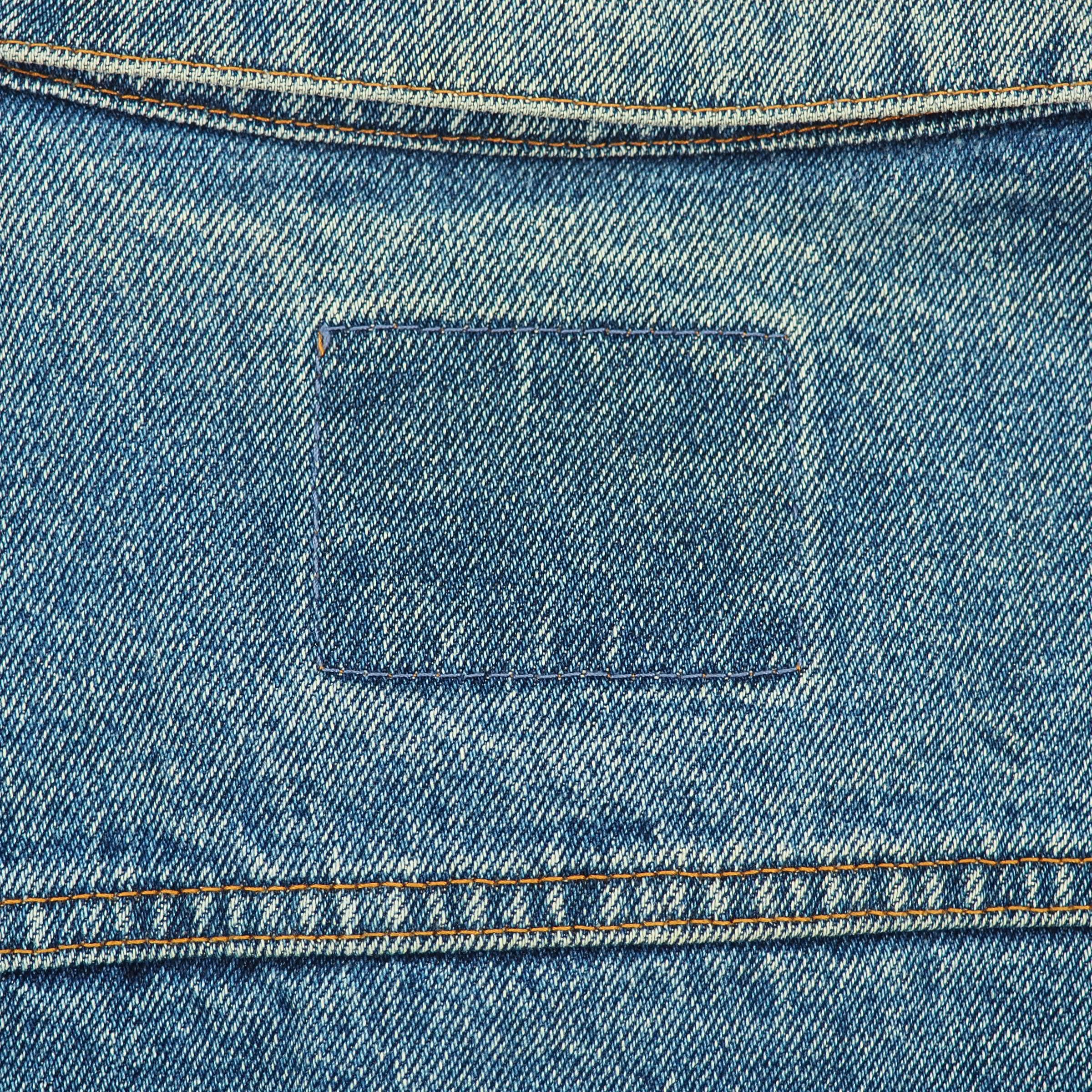 Close-up of a blue denim jacket showing the back pocket with a rectangular patch label and contrast orange stitching.