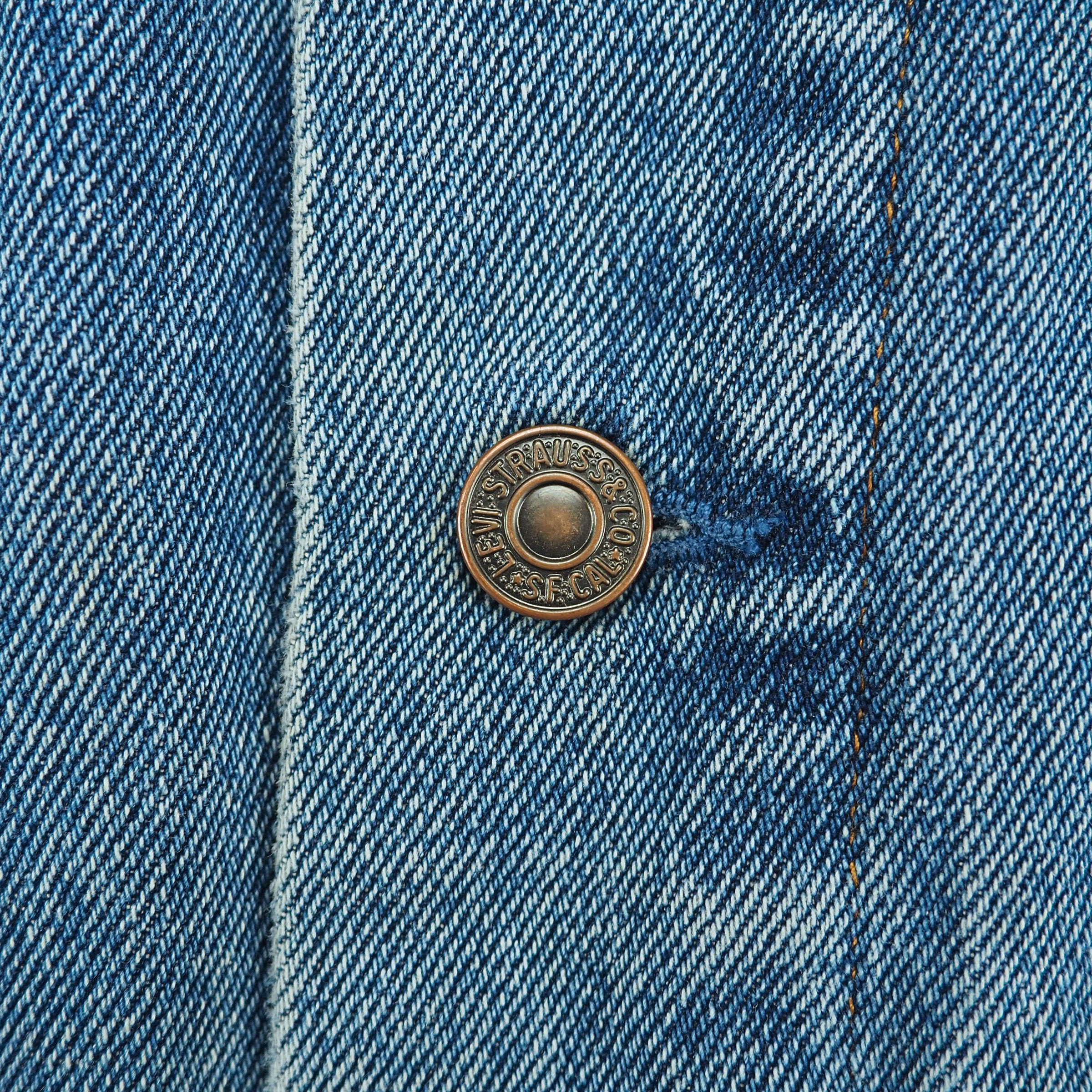 A close-up of a vintage-style bronze button on a blue denim jacket, showing the detailed twill texture of the fabric.