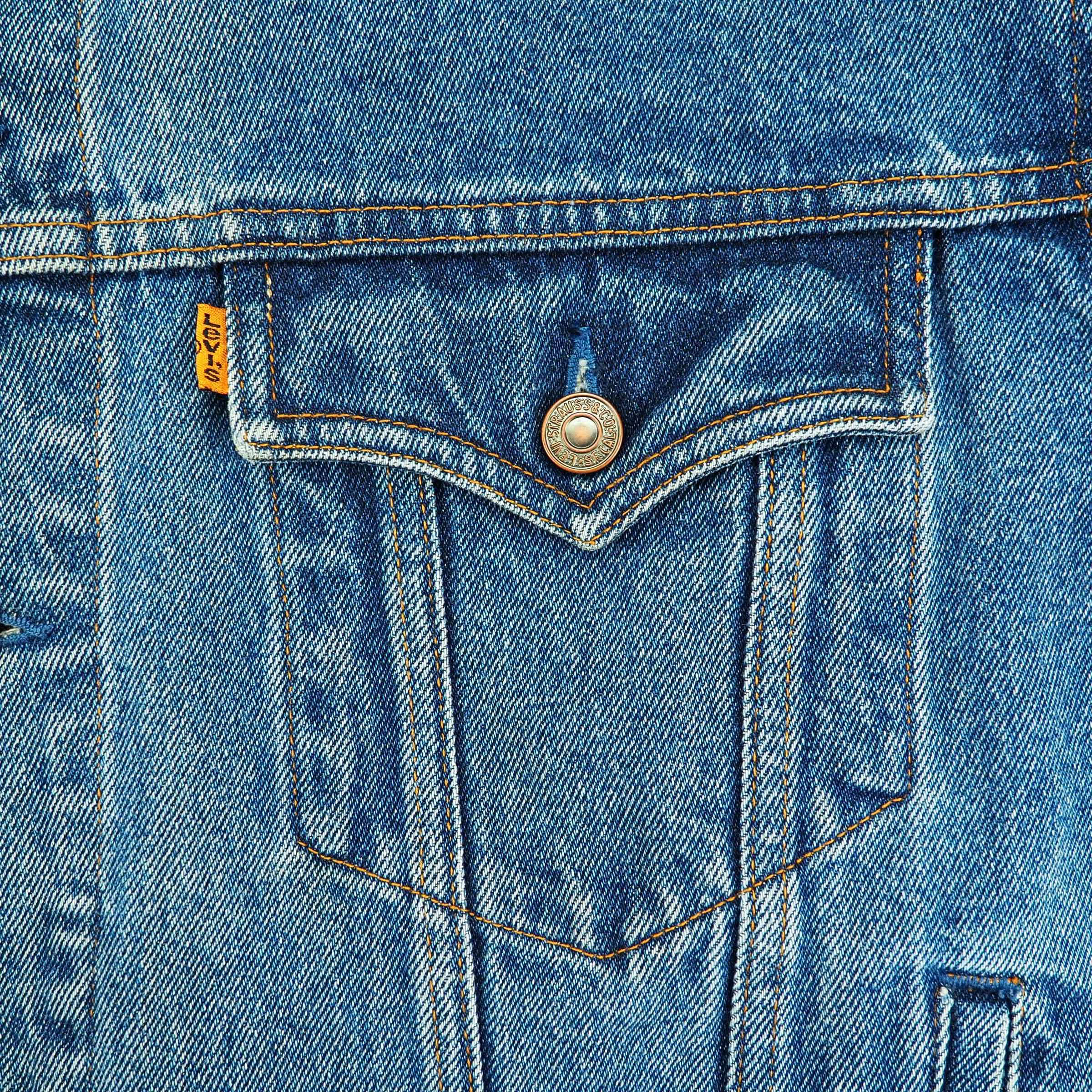 Close-up of a blue denim jacket showing a front flap pocket with a metal button, contrast stitching, and a small yellow Levi's brand tab.
