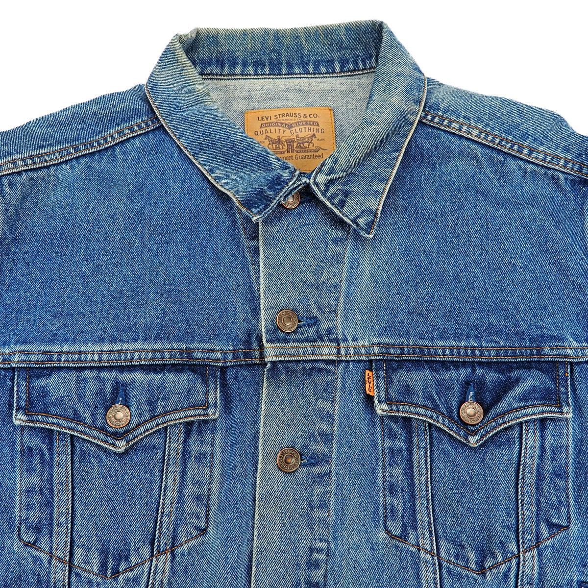 Close-up of a blue denim jacket with a pointed collar, button front, and two chest pockets. A tan leather patch with Levi's branding is visible inside the collar, and a small orange tab is on the right chest pocket.
