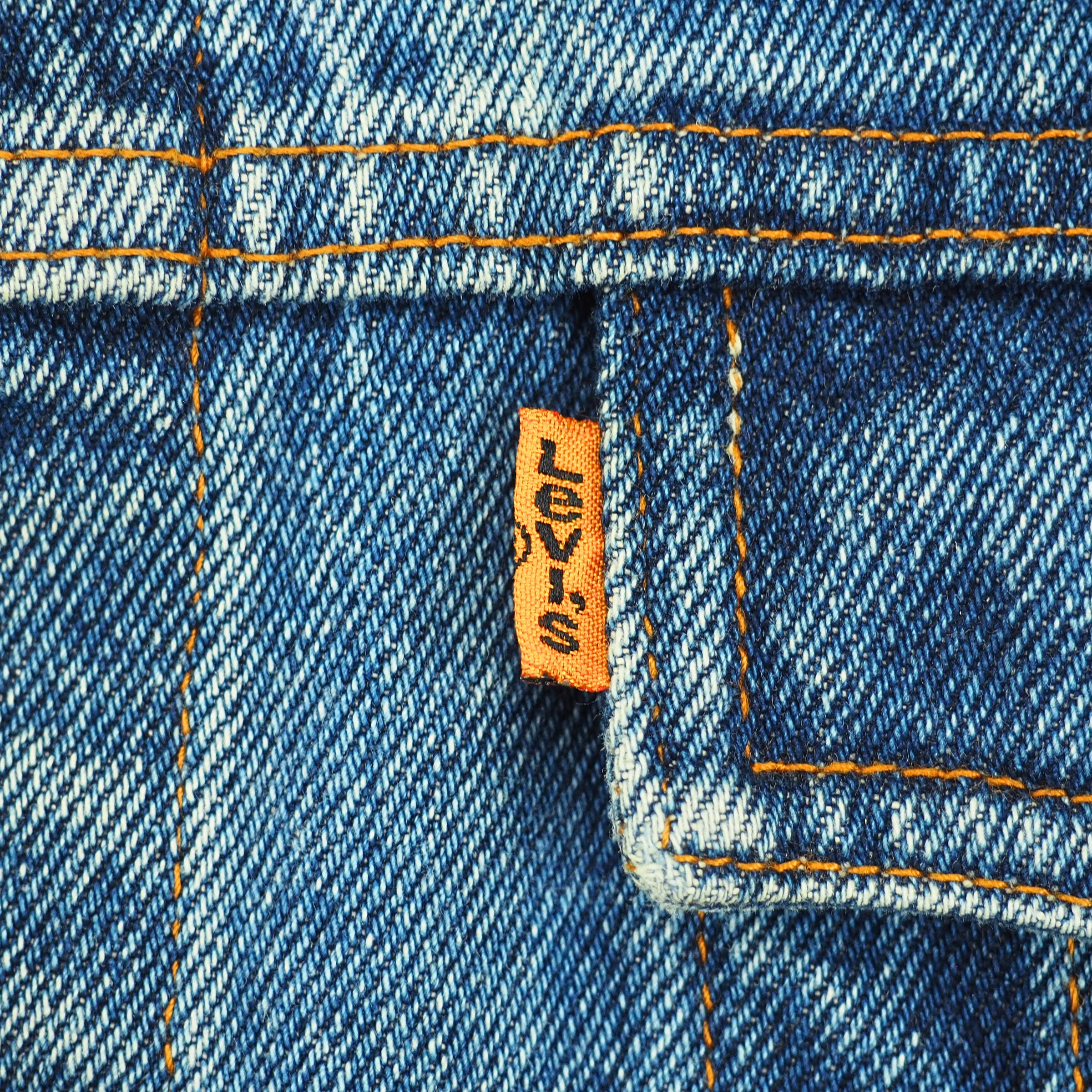 Close-up of blue denim fabric with orange contrast stitching and a small orange Levi's brand tab stitched onto the pocket.