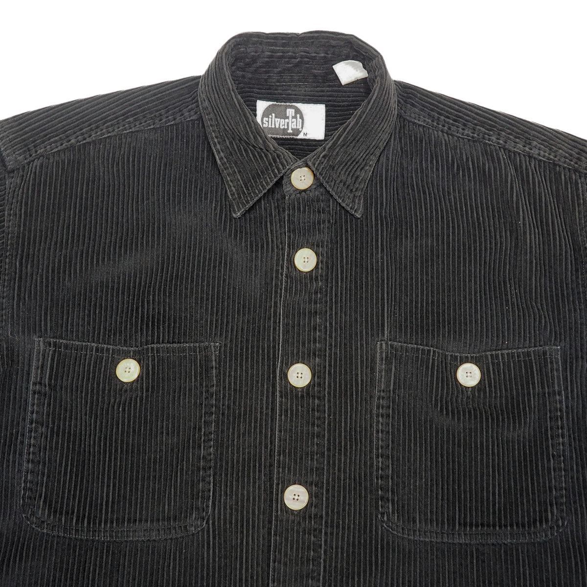 Close up of the chest and collar of a black corduroy button-down shirt jacket with two chest pockets and light-colored buttons, laid flat against a white background.