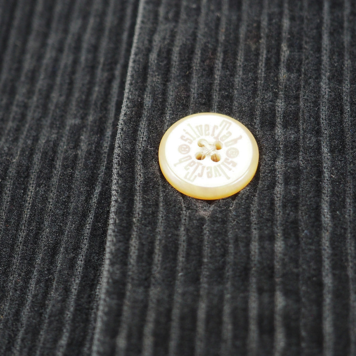 A close-up of a dark charcoal gray corduroy fabric with a single cream-colored button featuring the brand name 'SILVERTAB' printed on it.