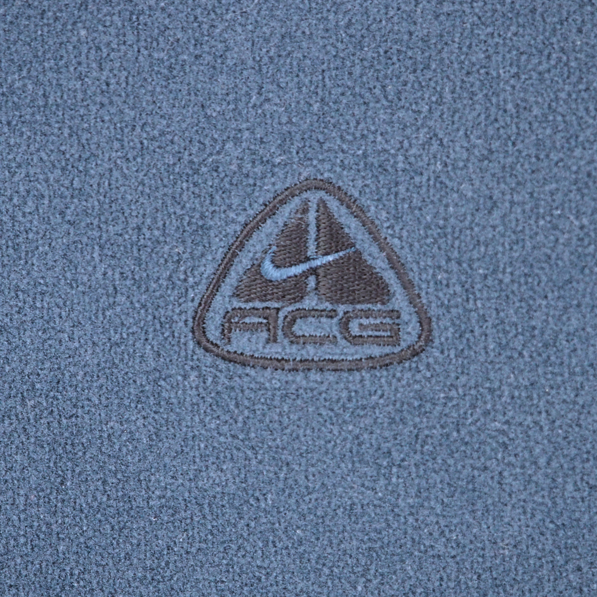 Close-up of a textured blue fabric with a central embroidered triangular logo featuring a swoosh and the letters ACG.