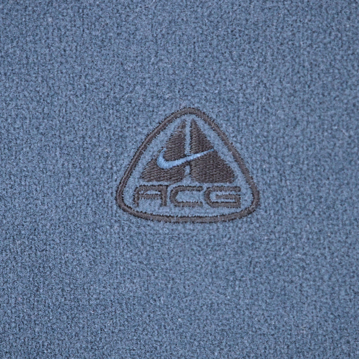 Close-up of a textured blue fabric with a central embroidered triangular logo featuring a swoosh and the letters ACG.