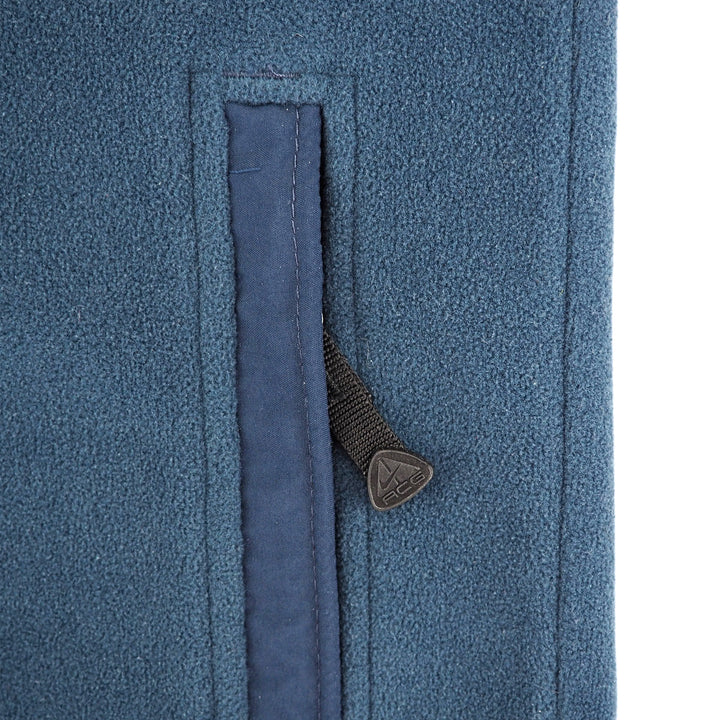 Close-up of a textured blue fleece jacket showing a vertical zippered pocket with a dark gray fabric pull tab featuring a triangular logo.