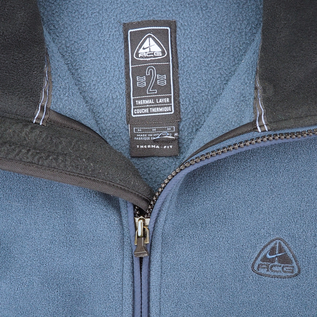 Close-up of a blue fleece zip-up jacket with dark gray trim, showing the inner neck label and a small chest logo of ACG.