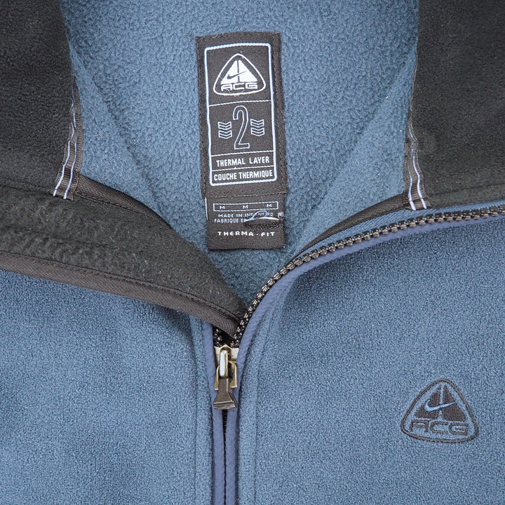 Close-up of a blue fleece zip-up jacket with dark gray trim, showing the inner neck label and a small chest logo of ACG.