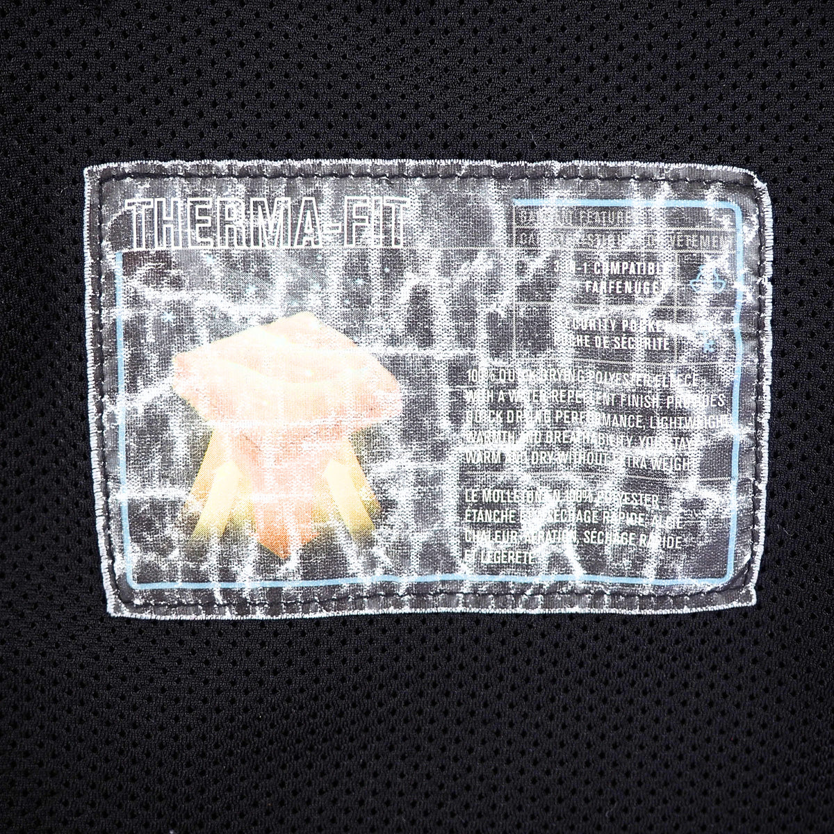 A close-up of a black mesh athletic garment showing a rectangular sewn patch with the text 'THERMA-FIT' and technical specifications, plus a small peach-and-yellow flower graphic.