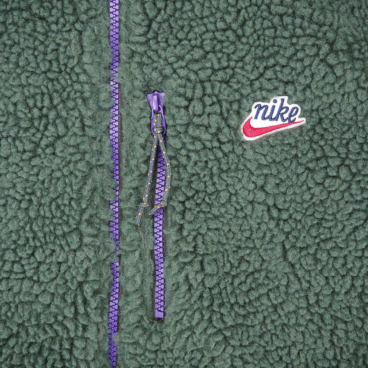 Close-up of a green sherpa fleece jacket featuring a purple zipper, a purple Nike logo tab, and a white and red embroidered Nike swoosh on the chest.