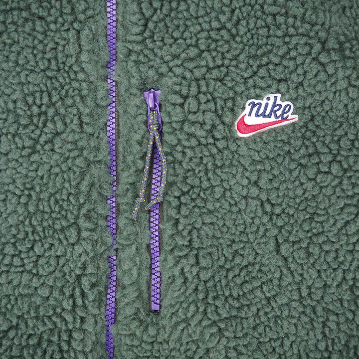 Close-up of a green sherpa fleece jacket featuring a purple zipper, a purple Nike logo tab, and a white and red embroidered Nike swoosh on the chest.