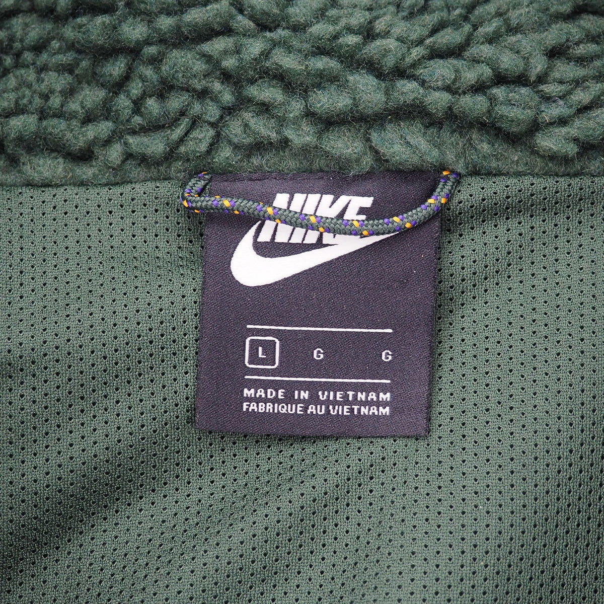 Close-up of a Nike garment tag sewn onto the inside of a dark green mesh fabric, with a chunky green sherpa lining above it. The tag shows the white Nike logo, size information, and 'MADE IN VIETNAM' text.