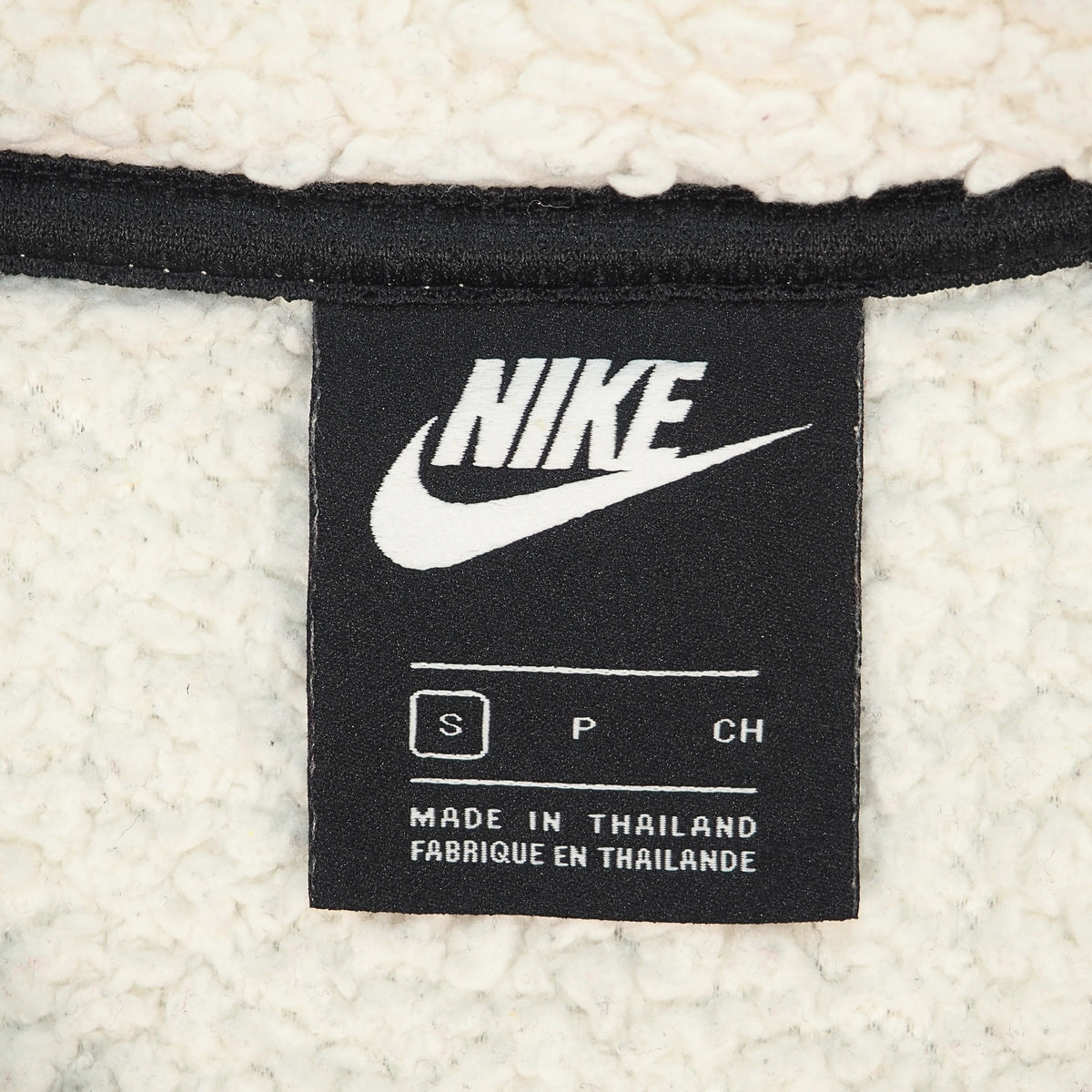 Close-up of a Nike brand tag sewn onto a cream-colored sherpa fleece garment. The black tag features the white Nike logo, size information (S, P, CH), and 'MADE IN THAILAND' text.