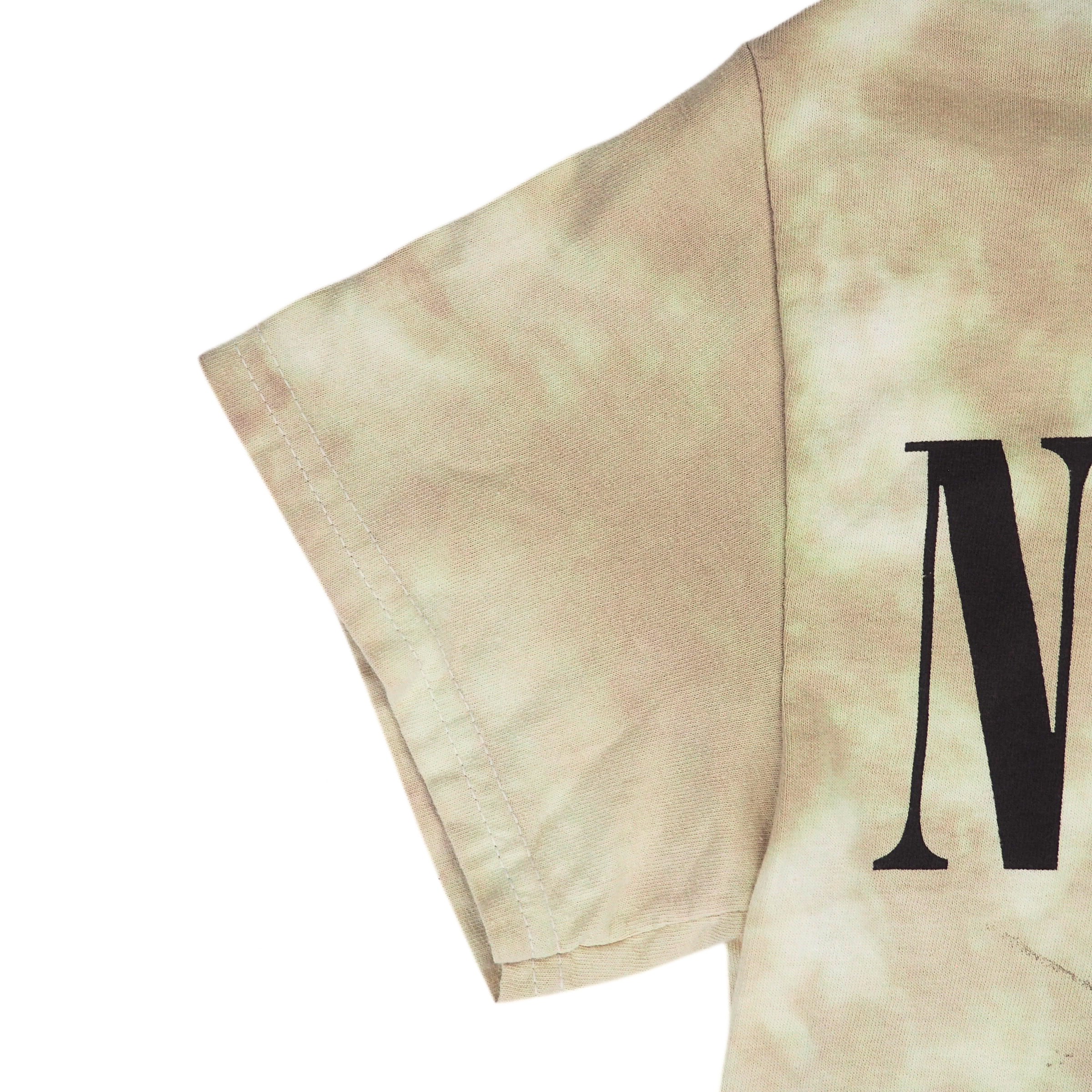 Close-up of a short-sleeve T-shirt sleeve showing a beige and off-white tie-dye pattern with a large black letter 'N' printed on the side, set against a plain white background.