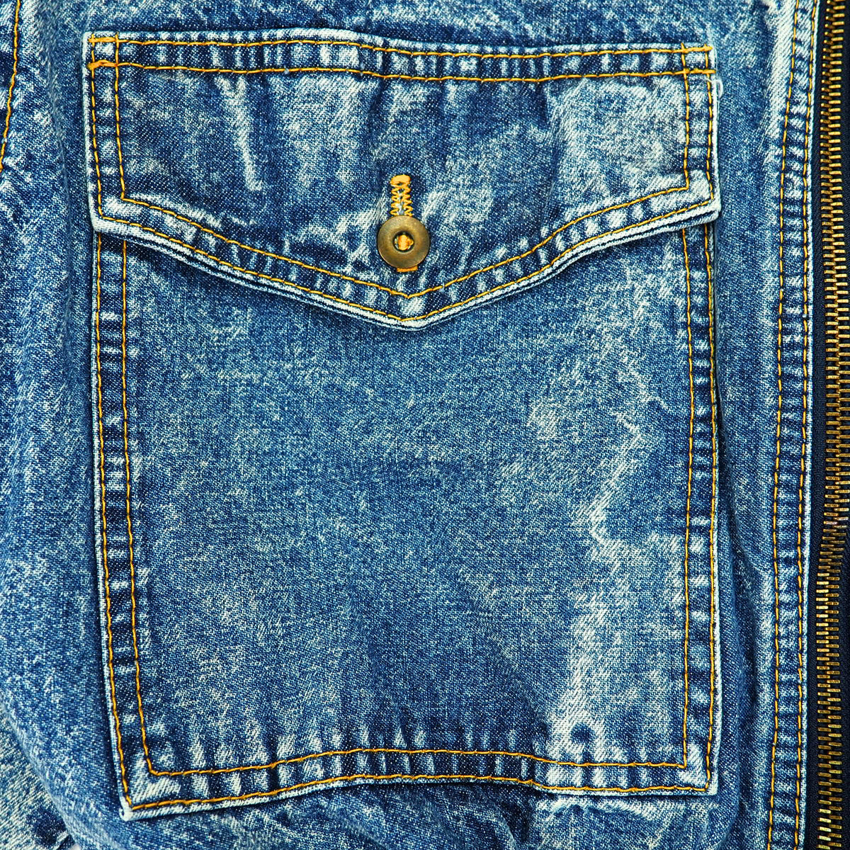 Close-up of a blue denim jacket featuring a front pocket with a gold-tone snap button and contrasting yellow-orange stitching.