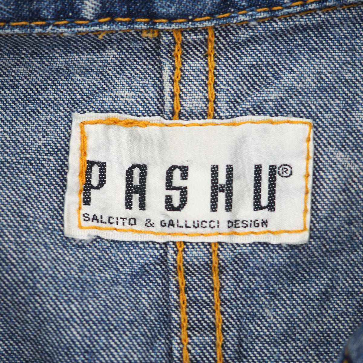 Close-up of a blue denim waistband with a white brand label reading PASHU and smaller text 'SALCITO & GALLUCCI DESIGN'. Yellow contrast stitching is visible around the label and down the center front.