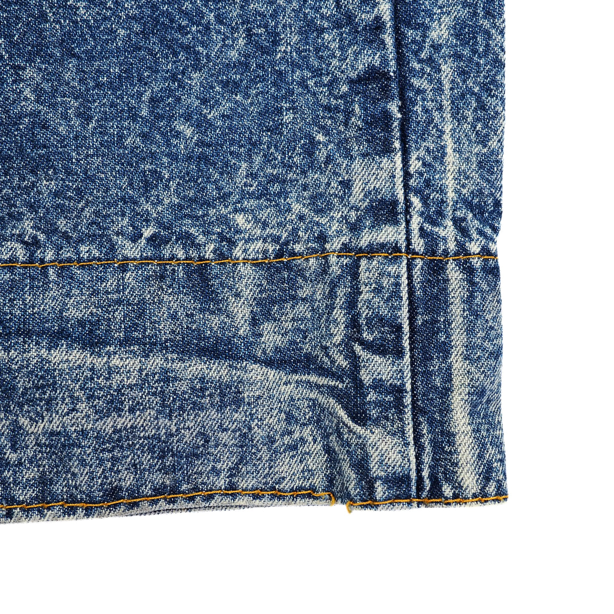 A close-up detail shot of a denim garment corner, showing the blue acid-wash texture, yellow contrast stitching, and raw hem against a white background.