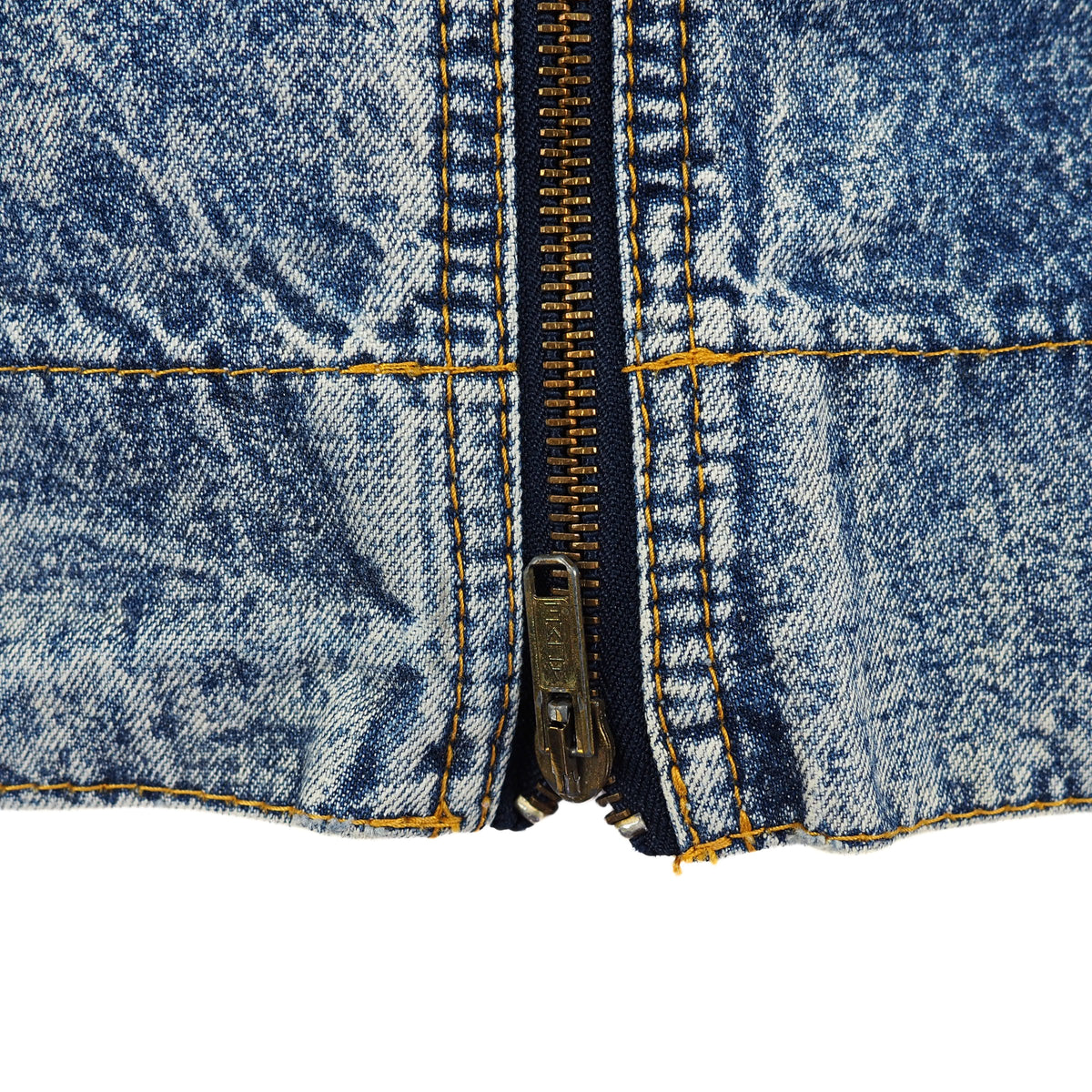 Close-up of a blue denim garment with a central metal zipper, yellow contrast stitching, and a white background visible at the bottom.
