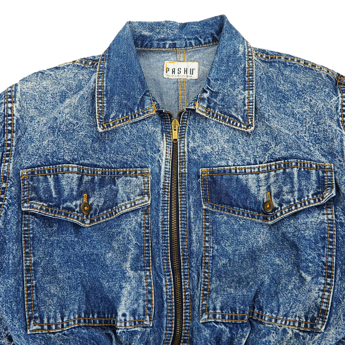 A close-up of a blue acid-wash denim jacket with a front zipper, two buttoned chest pockets, and a visible PASHMU brand label inside the collar.