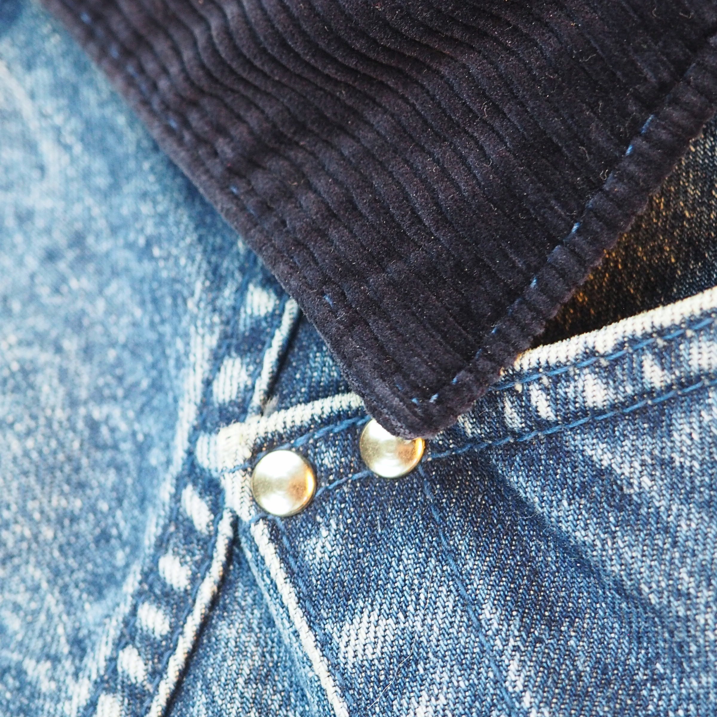 Close-up detail of a dark charcoal corduroy collar layered over blue denim fabric with metal snap buttons.