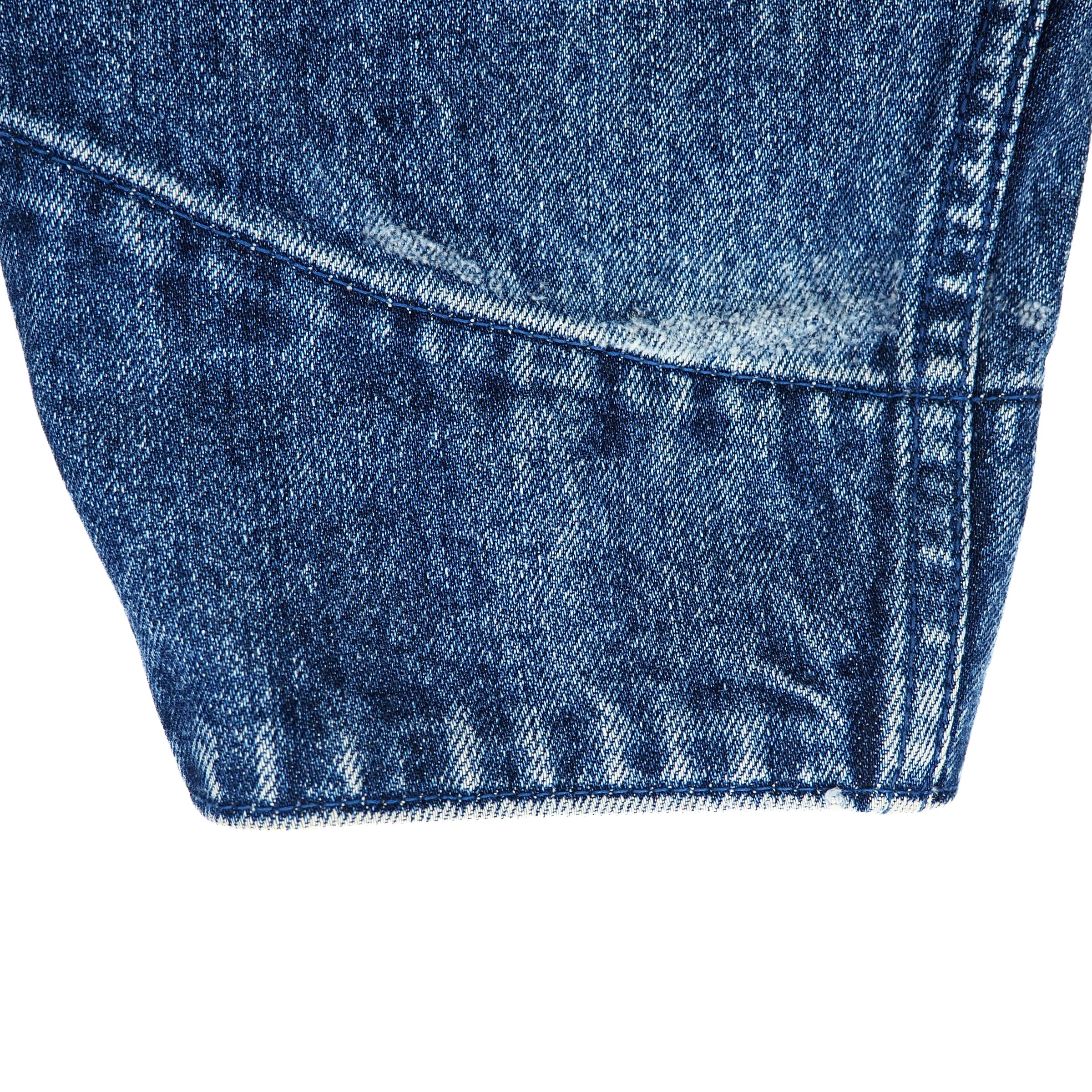 Close-up detail of a blue denim garment, showing the fabric texture, stitching, and a frayed hem edge against a plain white background.