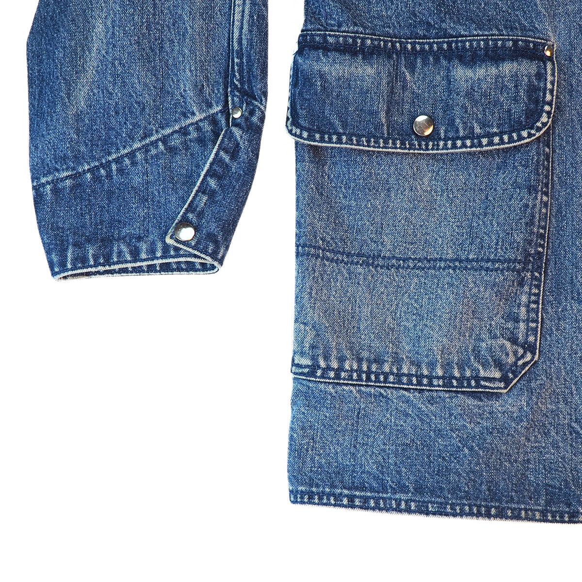Close-up detail of a blue denim jacket showing a buttoned flap pocket and a buttoned cuff on a white background.