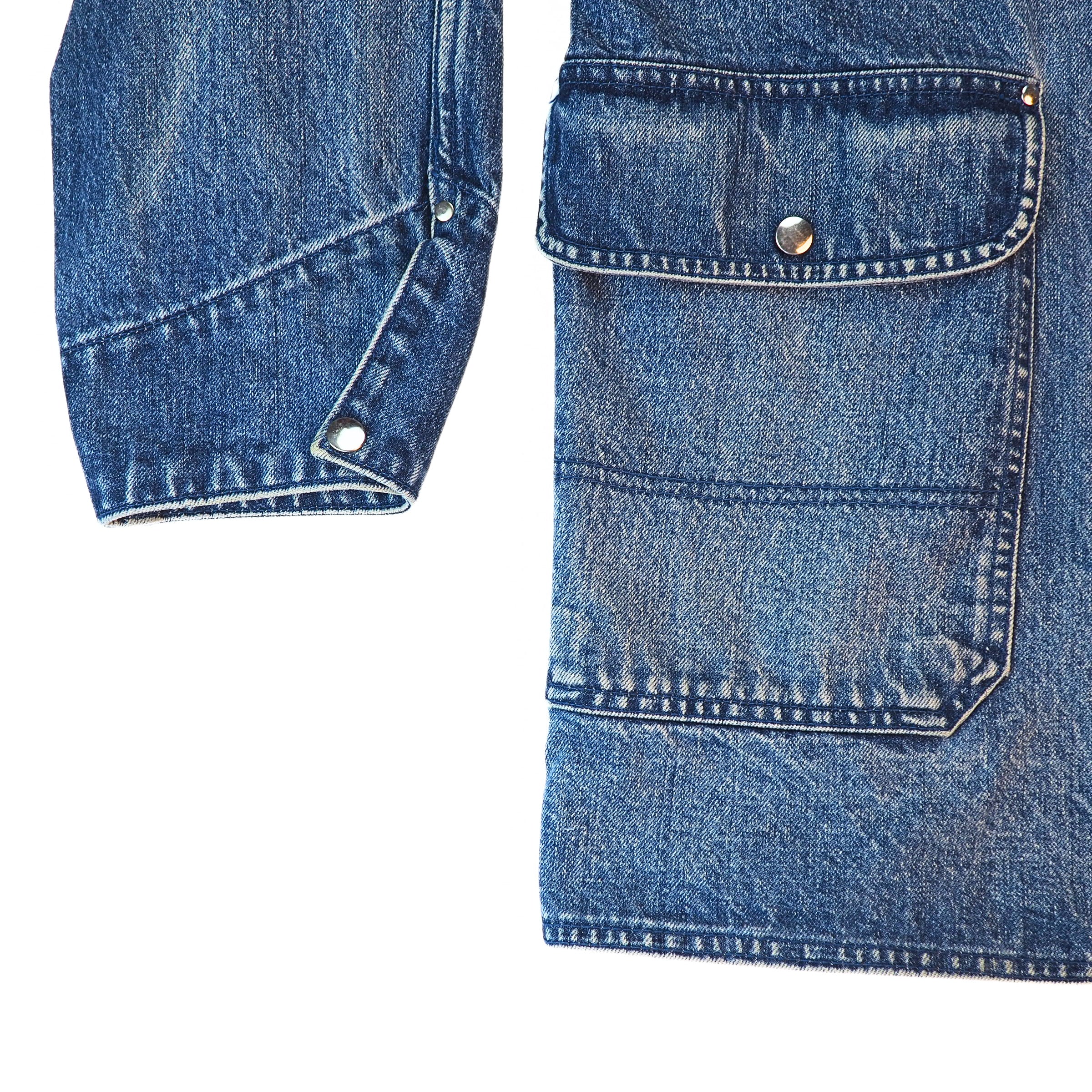 Close-up detail of a blue denim jacket showing a buttoned flap pocket and a buttoned cuff on a white background.