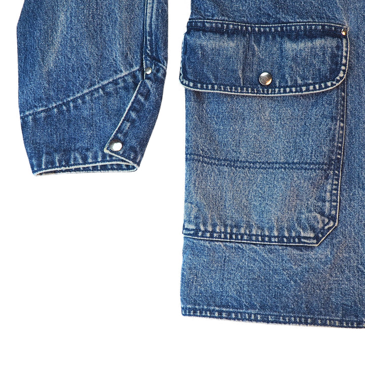 Close-up detail of a blue denim jacket showing a buttoned flap pocket and a buttoned cuff on a white background.