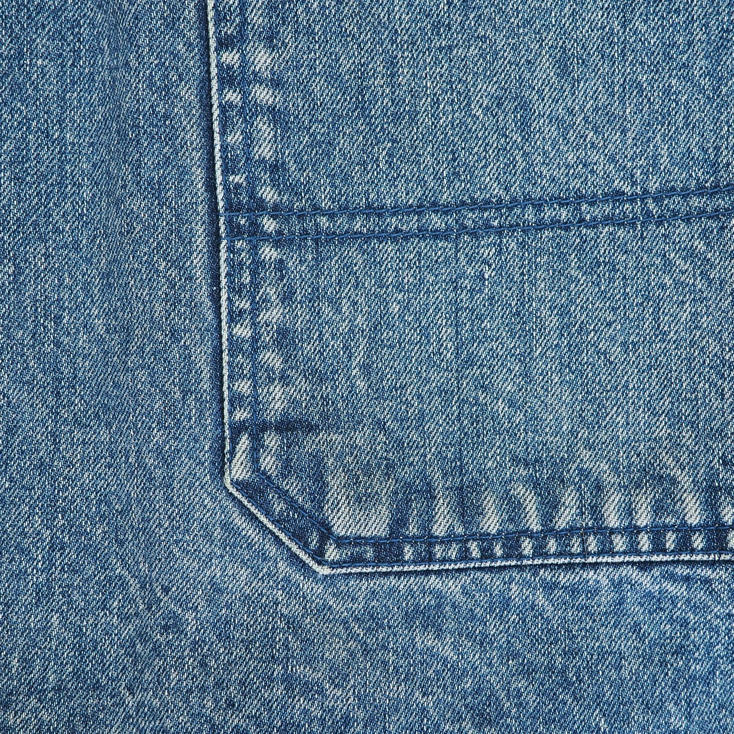 Close-up of a blue denim fabric texture showing the corner of a pocket with detailed stitching.