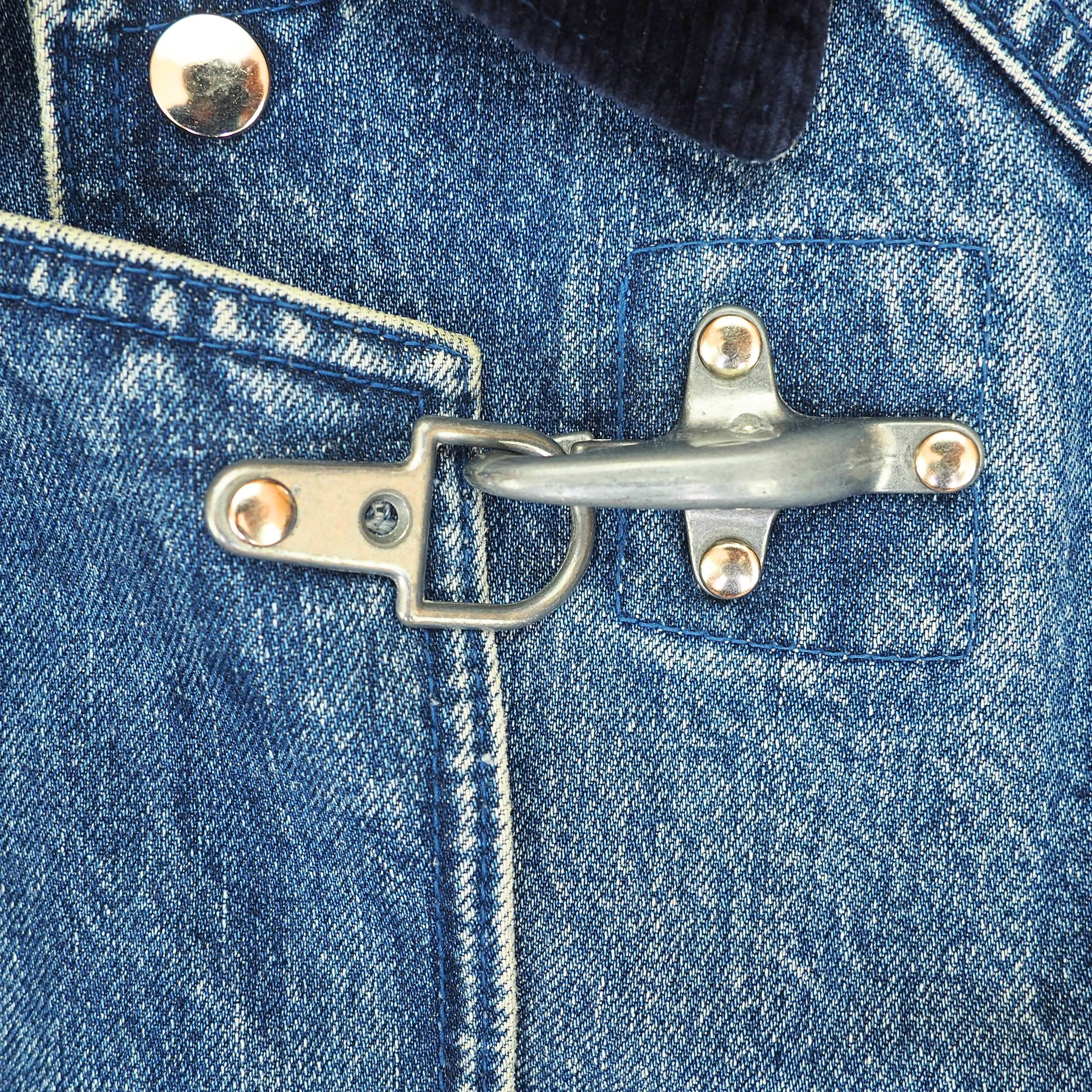 A close-up of a blue denim jacket featuring a unique silver metal toggle clasp closure on the chest pocket, with visible rivets and stitching.