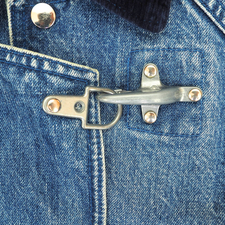A close-up of a blue denim jacket featuring a unique silver metal toggle clasp closure on the chest pocket, with visible rivets and stitching.
