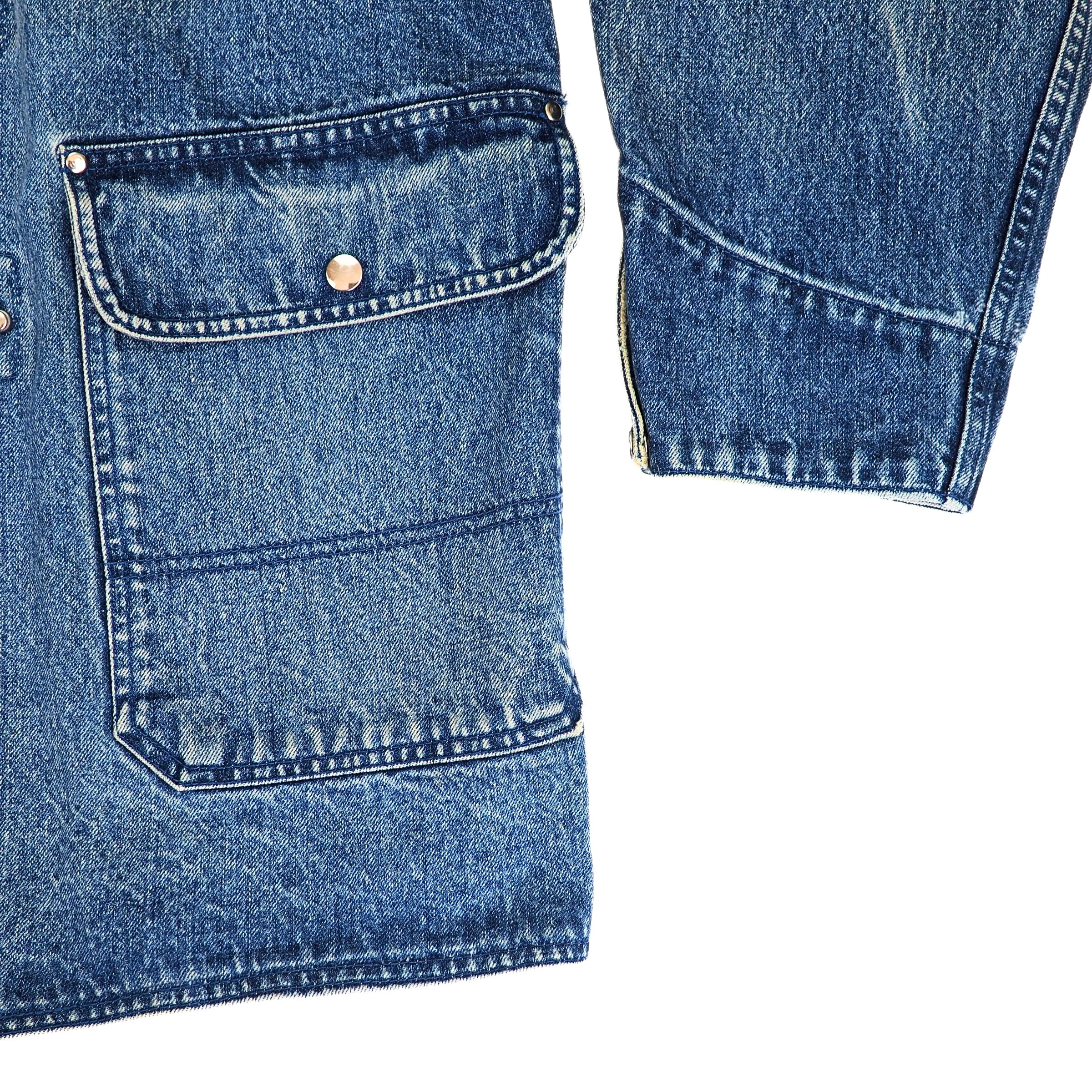 Close-up detail of a blue denim garment showing a front flap pocket with a brass rivet and a zippered cuff on a white background.