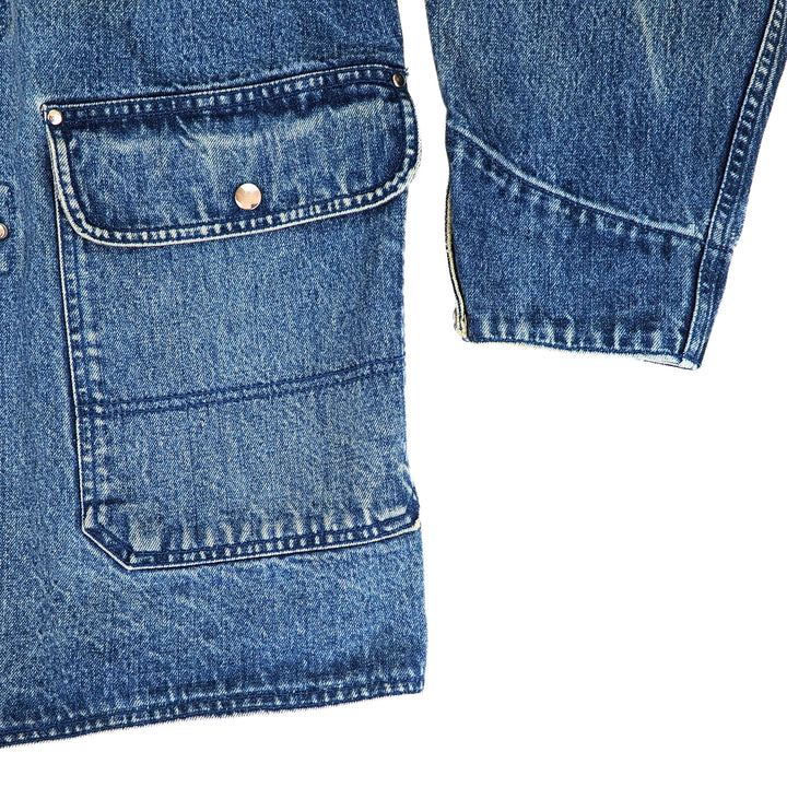 Close-up detail of a blue denim garment showing a front flap pocket with a brass rivet and a zippered cuff on a white background.