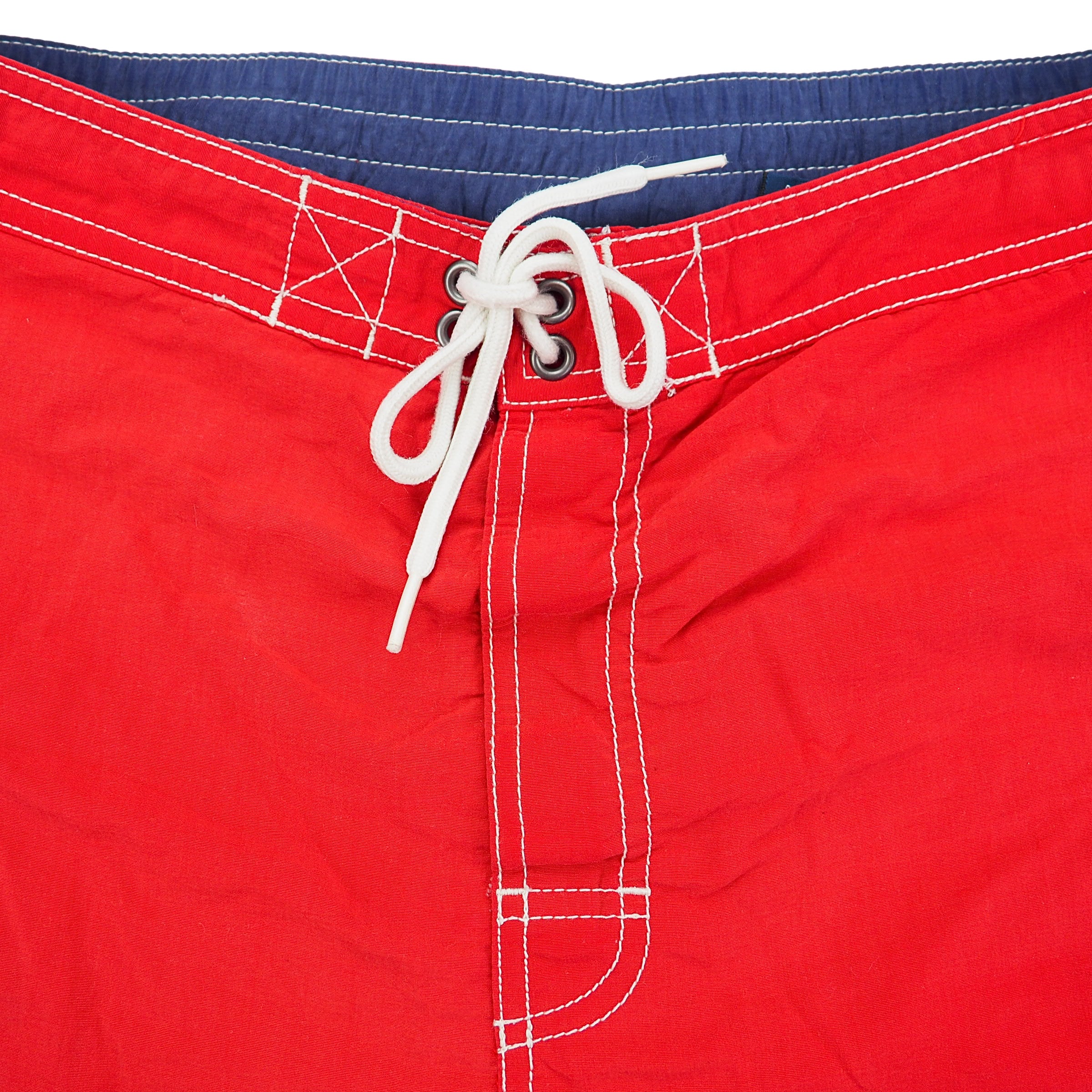 Red swim shorts with white drawstring and blue waistband on a white background