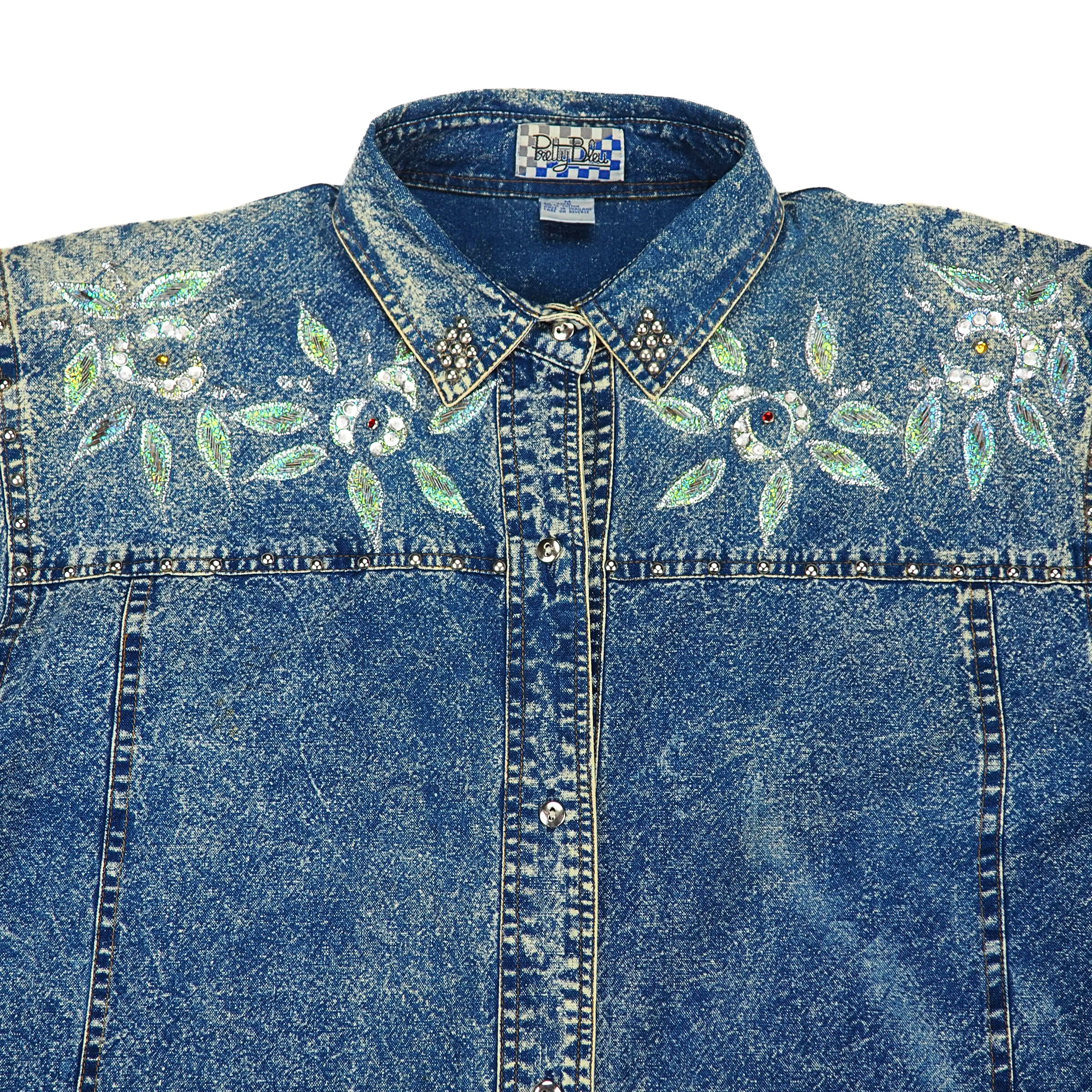 Western style 90s acid wash blue denim shirt with floral embroidery and decorative silver buttons on a white background