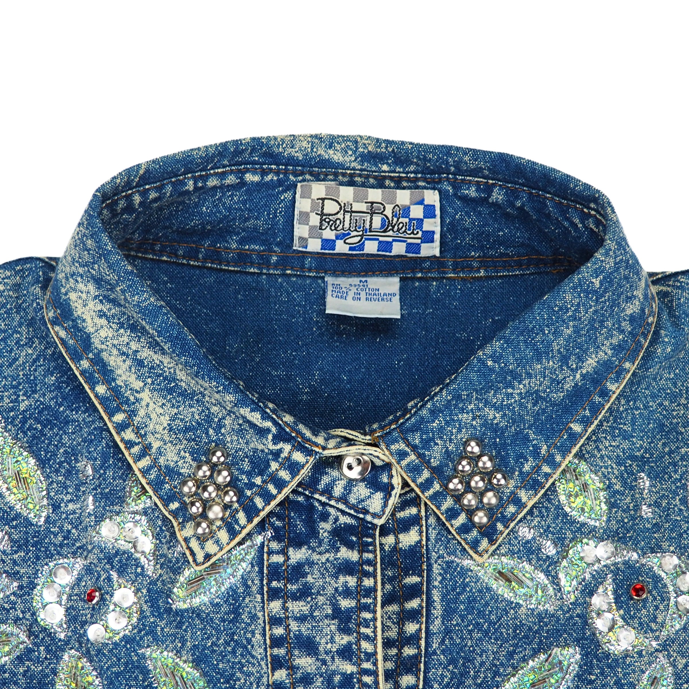 Close-up of a blue acid wash denim shirt with floral embroidery, beading, decorative buttons and a 'Pretty Bleu'  brand label inside the collar.