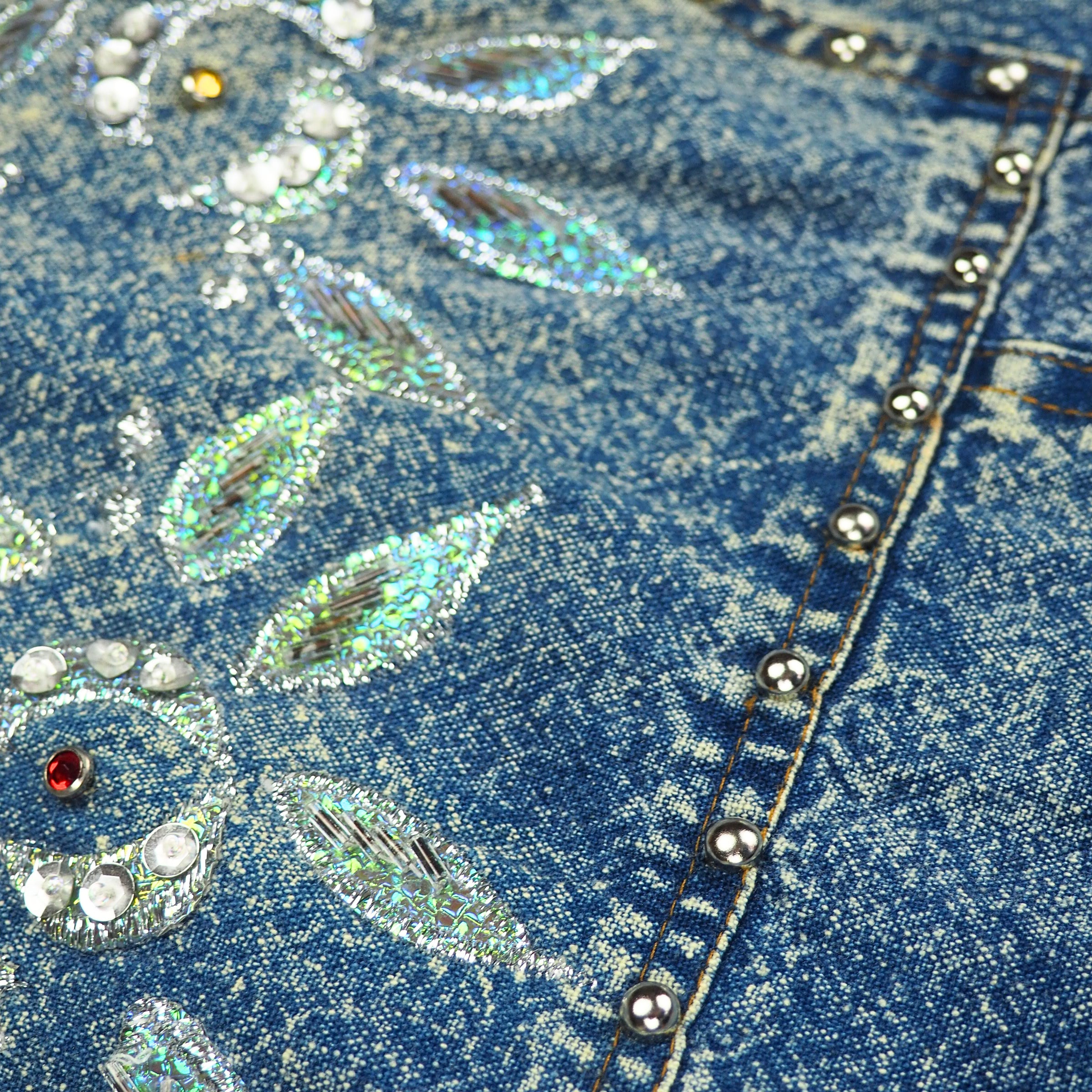 Close-up of blue acid wash denim fabric with decorative floral stitching and beading.